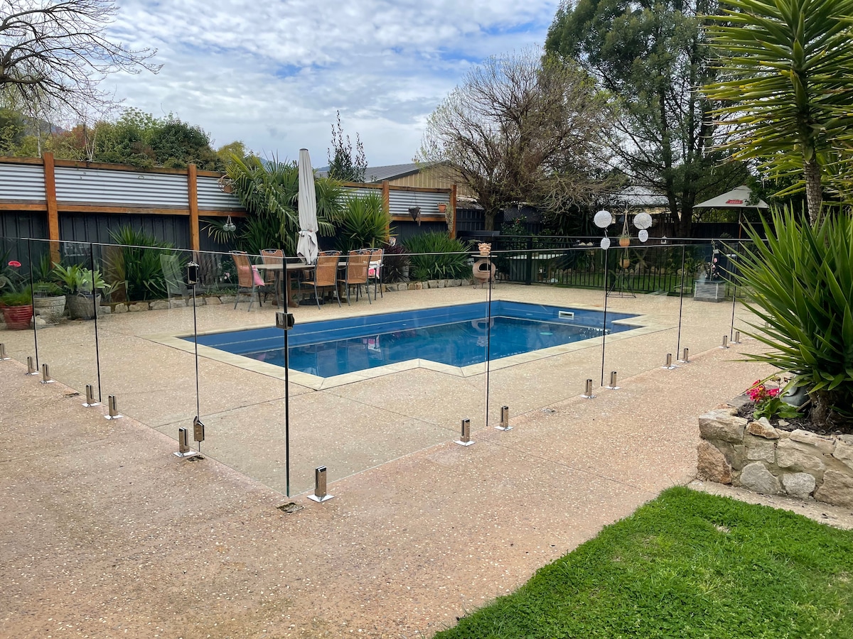 A fenced pool area is depicted with a clear glass barrier surrounding a rectangular pool. Comfortable outdoor seating with umbrellas can be seen nearby, along with landscaped greenery and decorative plants, providing a pleasant outdoor environment.