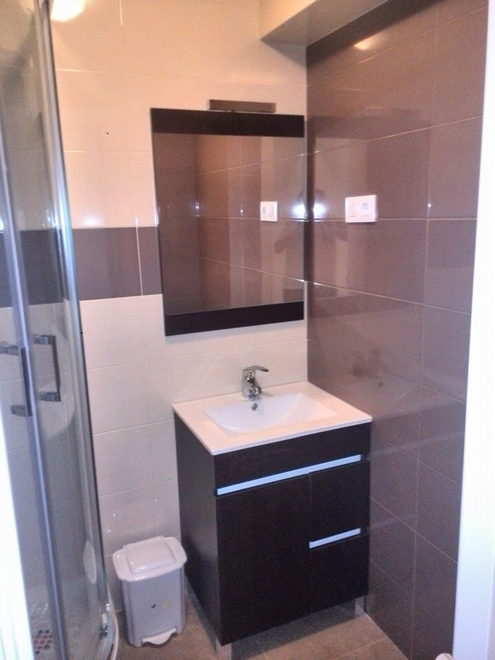 A compact bathroom features a modern vanity with a single sink and storage drawers. A glass shower enclosure is visible, alongside a small trash can. Wall tiles in varying shades of brown create a subtle contrast with the light-colored tiles on the floor.