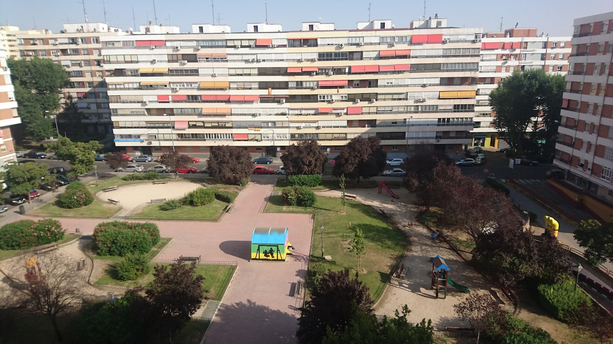 A view of a public park is visible, featuring well-maintained paths and greenery. Several multi-story residential buildings surround the area, showcasing a mix of colors and designs. Play areas with structures for children are present, along with shaded seating and pathways for leisurely strolls.