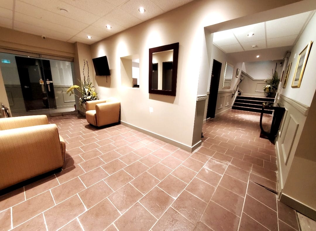 A spacious lobby area is furnished with two comfortable tan armchairs. A large mirror reflects the soft lighting above, while modern artwork adorns the walls. The tiled floor and subtle decorative plants contribute to a welcoming atmosphere, leading towards the staircase in the background.