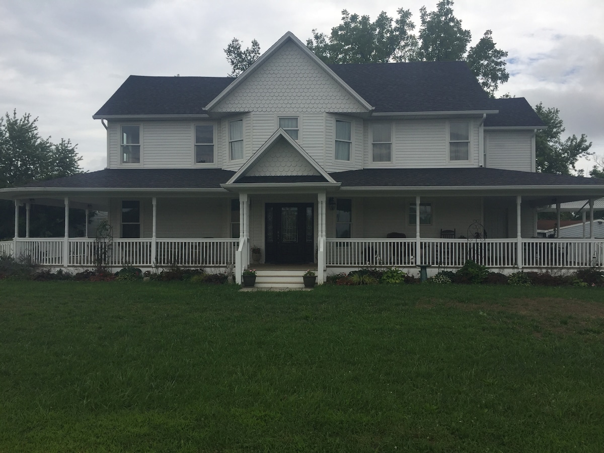 The historic Gourley Manor features a two-story white exterior, showcasing multiple gabled roofs and a wraparound porch with white railings. Landscaping includes grass and flower beds, while large windows provide a glimpse of the home's interior. Trees are visible in the background.