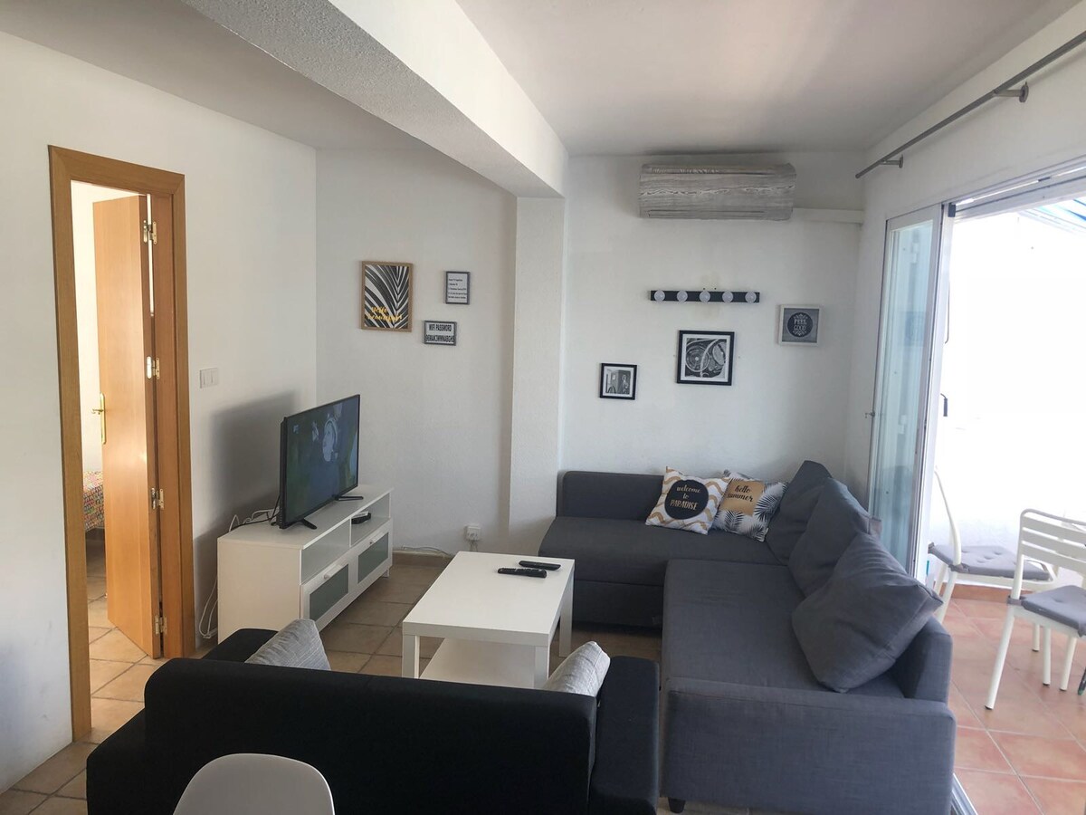 A bright living area is presented with two sofas in contrasting colors and a central coffee table. A television is situated on a media unit, while wall art and framed pictures add subtle character. Large glass doors lead to a terrace, enhancing natural light.
