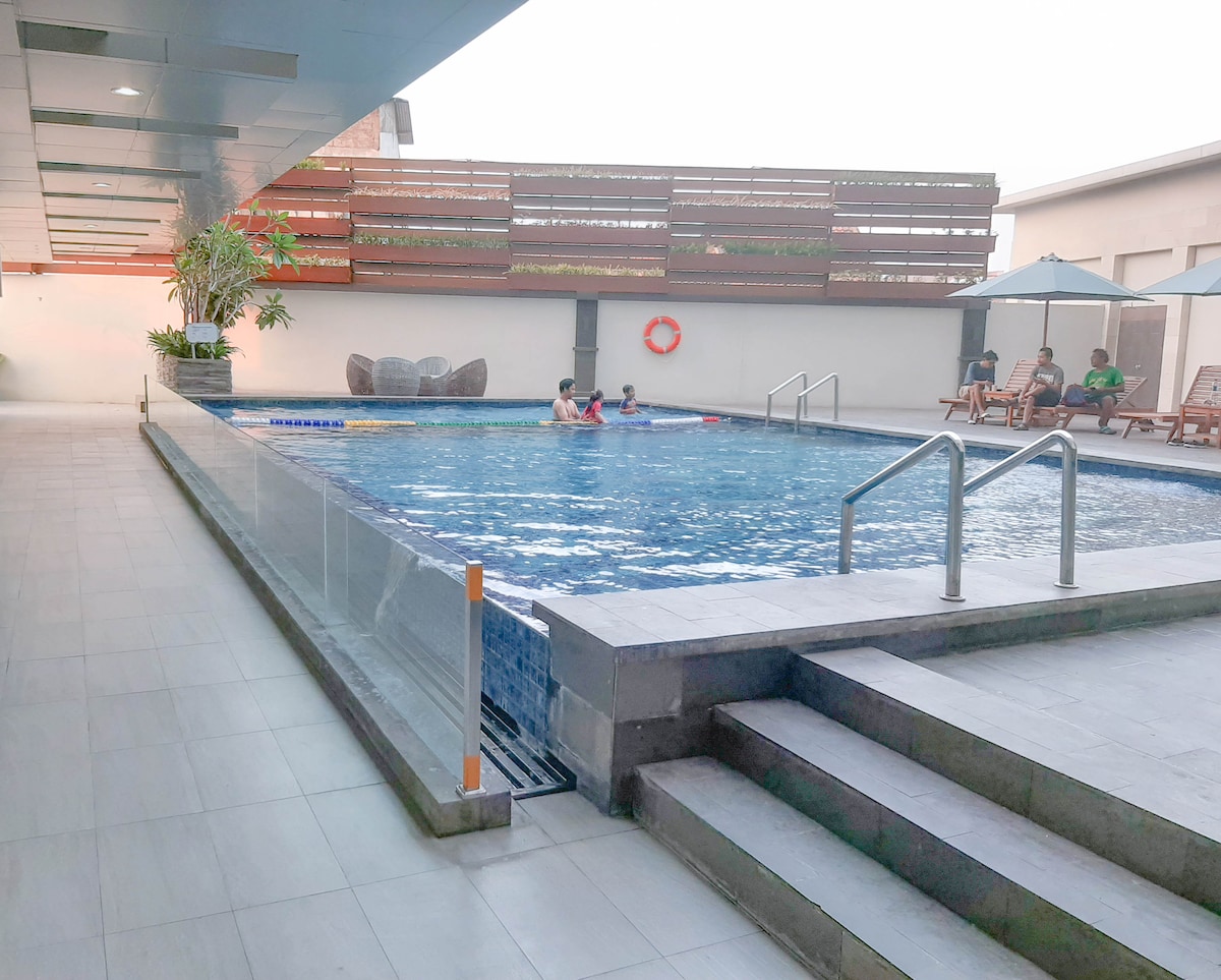 A swimming pool area is shown, featuring clear water and steps leading into it. Lounge chairs are positioned nearby, alongside umbrellas for shade. Lush greenery is visible in the background, with some guests enjoying the water, creating a relaxed atmosphere.