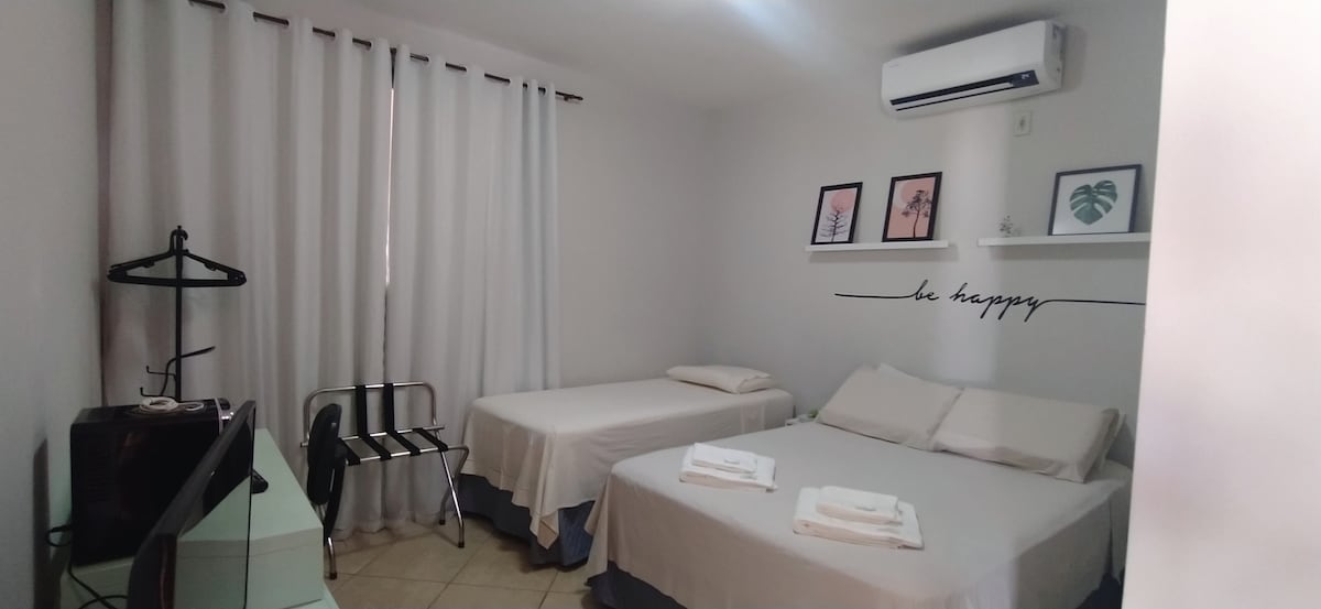 A bright and airy room is shown, featuring two spacious beds dressed in simple white linens. A small desk with a chair and a Smart TV is visible, along with a clothing rack. Soft natural light filters through the white curtains.
