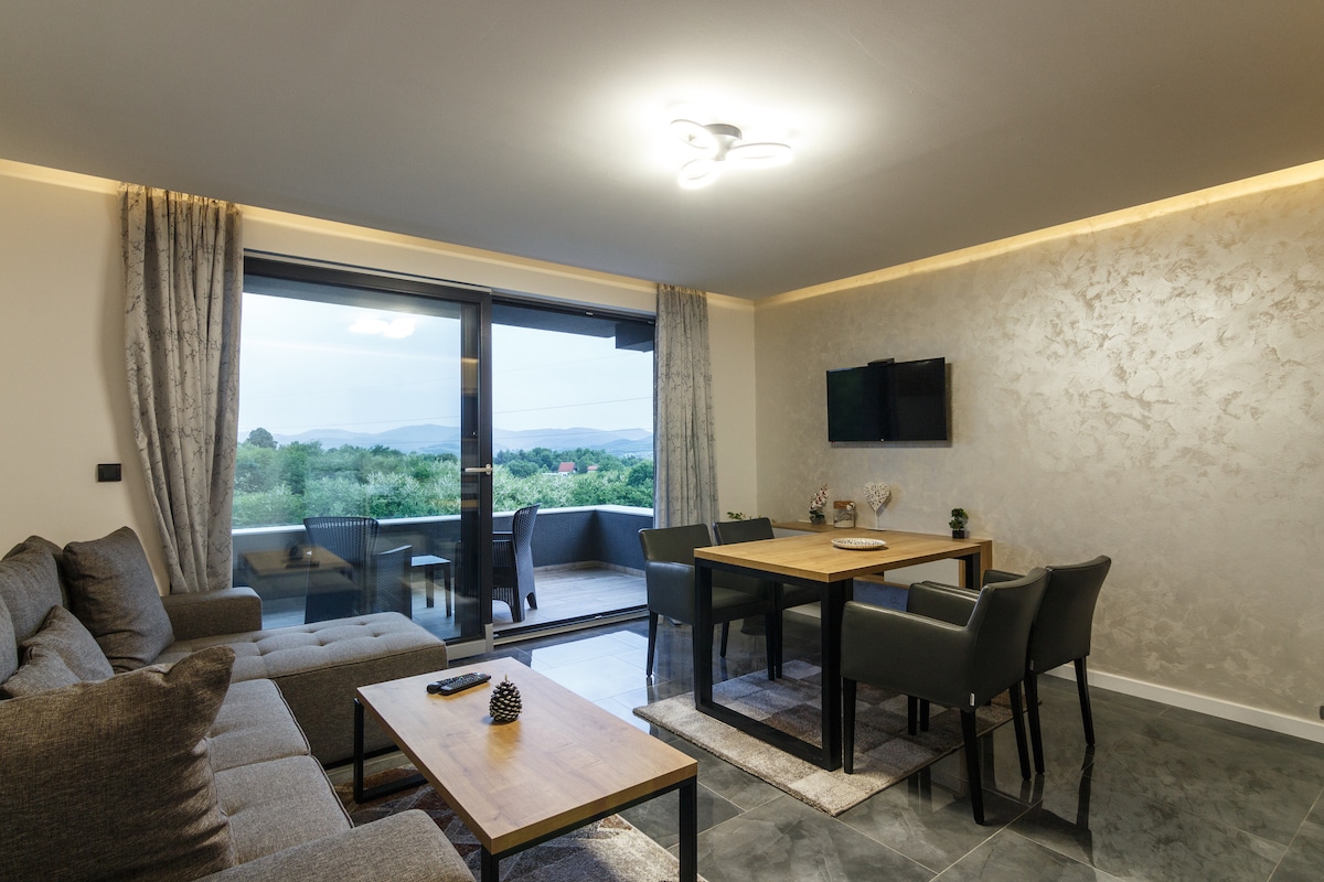 A modern living room showcases a comfortable grey sectional sofa and a wooden dining table surrounded by dark chairs. A flat-screen TV is mounted on a decorative wall, and large sliding glass doors lead to a terrace with green views beyond.