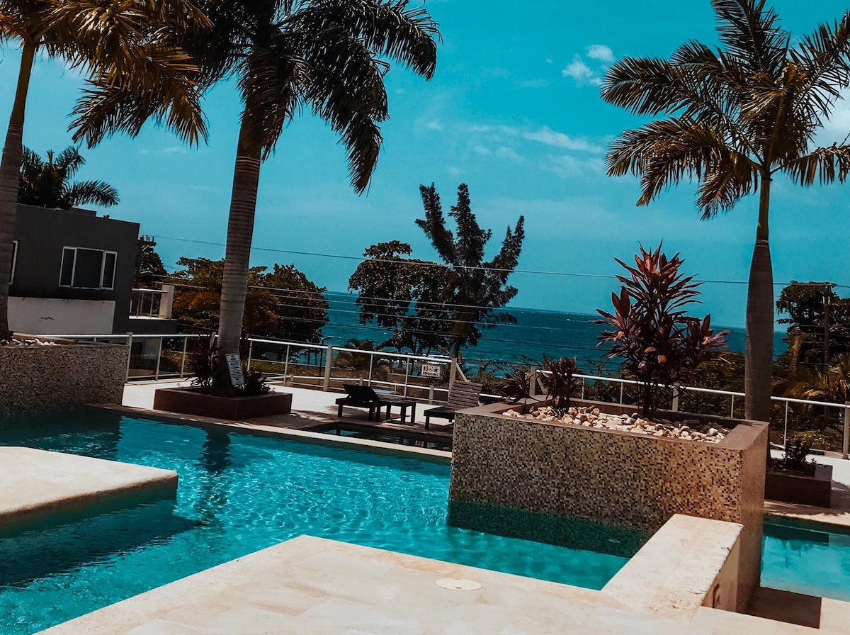 A tranquil pool area is framed by tall palm trees, offering views of the ocean in the background. The pool features smooth stone edges and lounge chairs are positioned nearby, inviting relaxation amid a serene tropical setting.