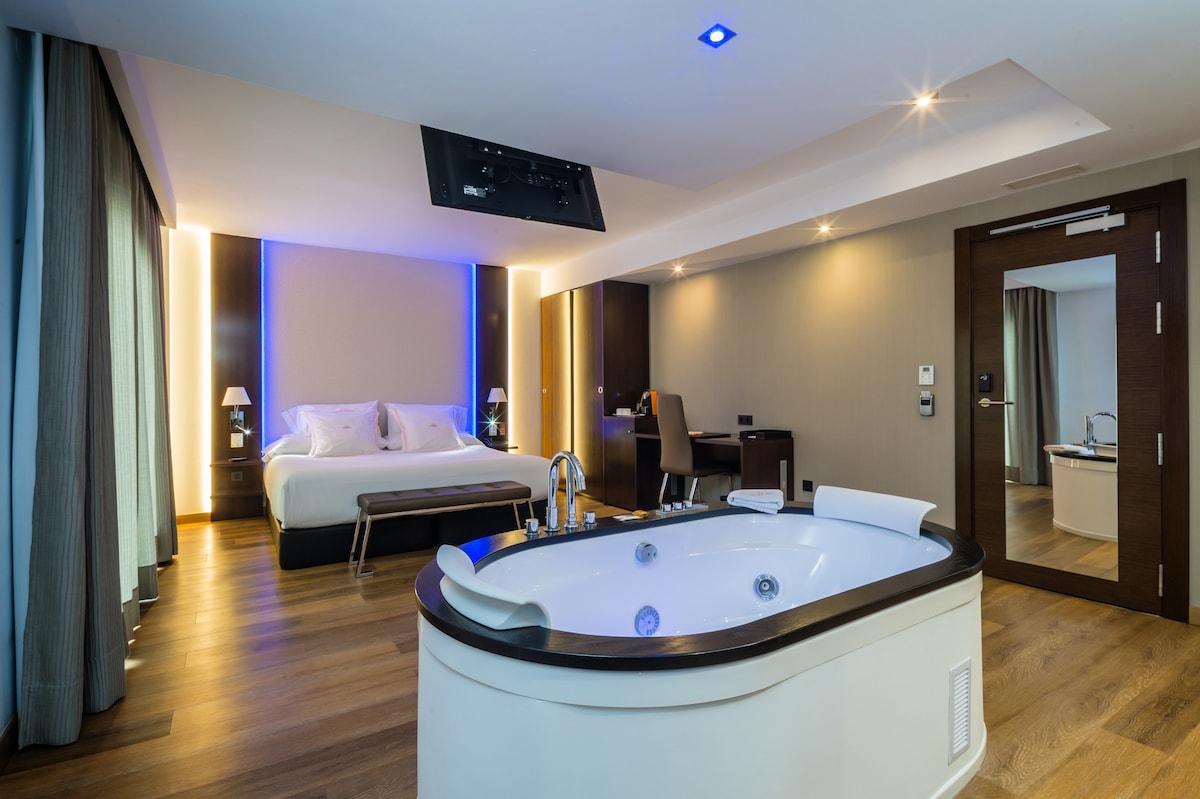 A modern hotel room features a spacious layout with warm wooden flooring. A king bed is positioned against a softly illuminated wall. A luxurious hydromassage tub is present, alongside a sleek television mounted on the ceiling, complementing the stylish decor.