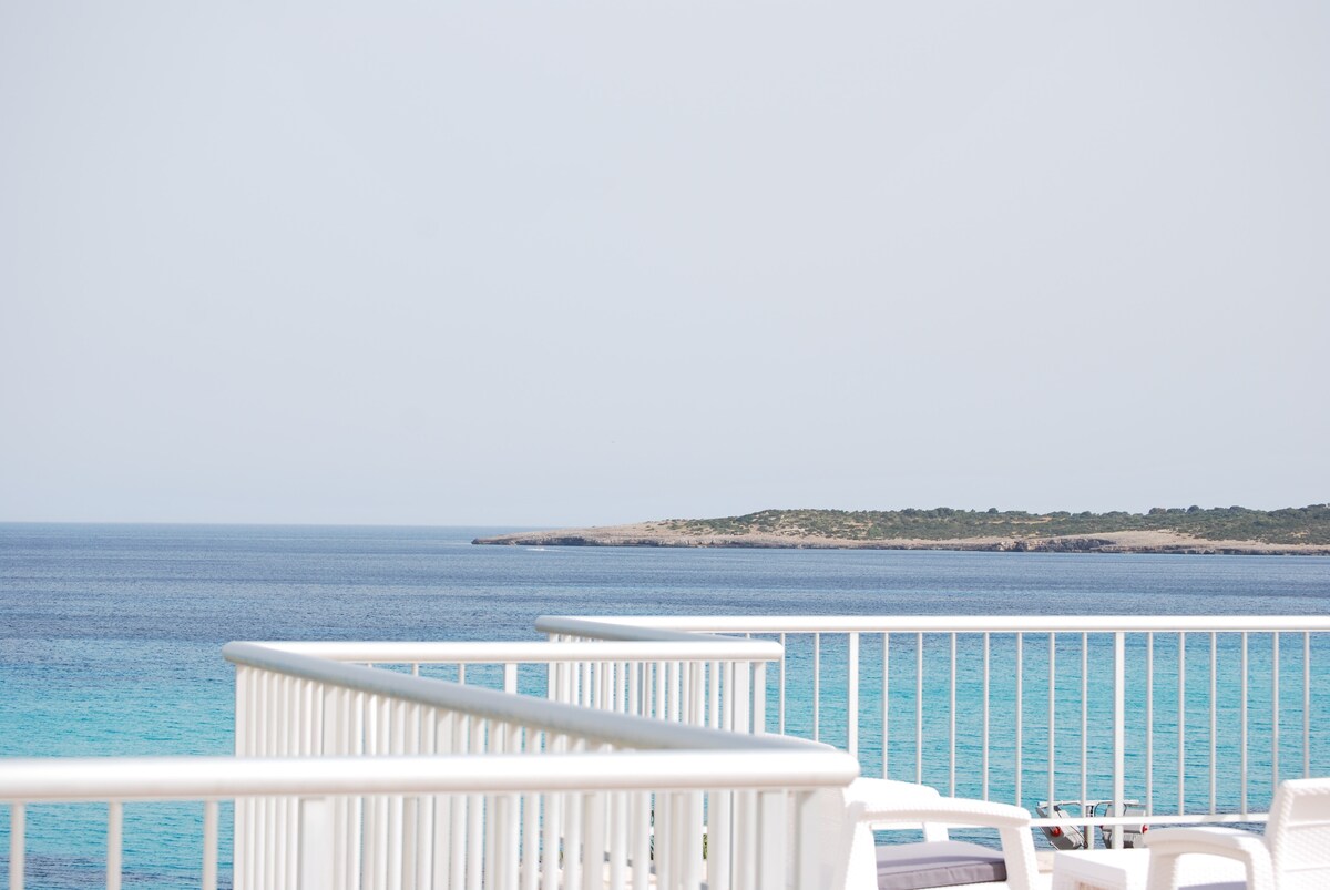 A tranquil view of the Mediterranean Sea is showcased, featuring varying shades of blue. The horizon is marked by a gentle shoreline, with a distant landmass visible. The image includes a white railing, highlighting the outdoor terrace space.