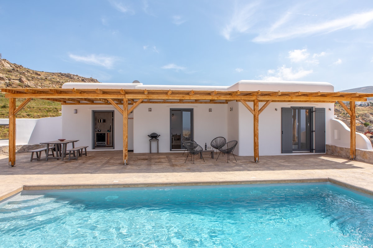 The villa features a private pool surrounded by a spacious terrace. An outdoor dining area with a wooden table is visible, complemented by several colorful chairs. Large sliding doors provide access to the interior while an overhead pergola offers shade, enhancing outdoor comfort.