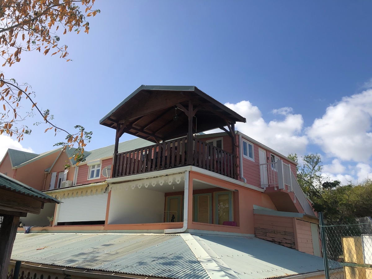 A two-story residence is shown, featuring a covered upper terrace with wooden beams, offering shaded outdoor space. The exterior walls are painted in soft tones, and multiple windows are visible, allowing natural light. A metal roof adds to the overall structure, enhancing the inviting feel.