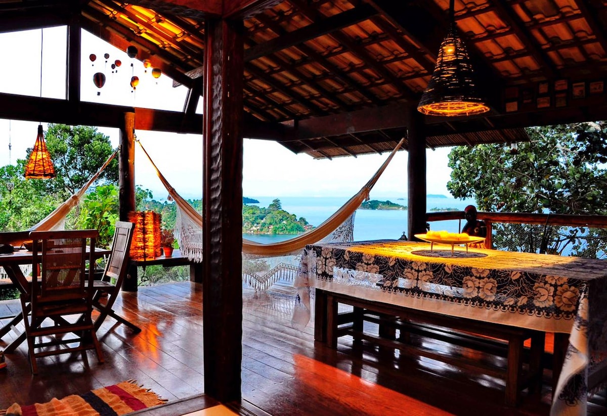 An open and airy living space features wooden beams and large windows offering views of the surrounding trees and the bay. A dining table is set near the window, while hammocks provide a relaxing spot. Warm lighting enhances the inviting atmosphere.