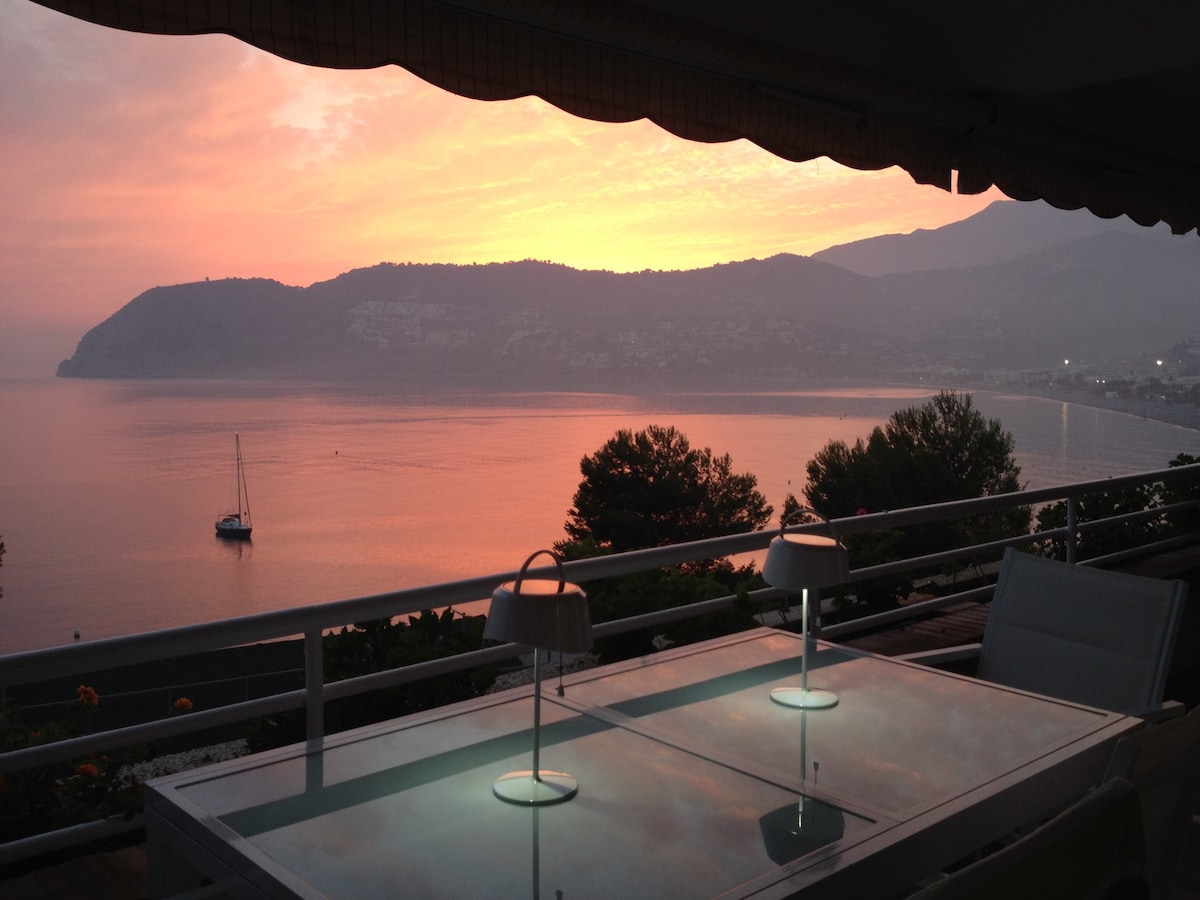 A spacious terrace is visible, featuring a glass table illuminated by stylish lamps. The view showcases a serene bay at sunset, with shades of pink and orange reflecting on the water. A sailboat rests peacefully in the foreground, framed by distant hills.