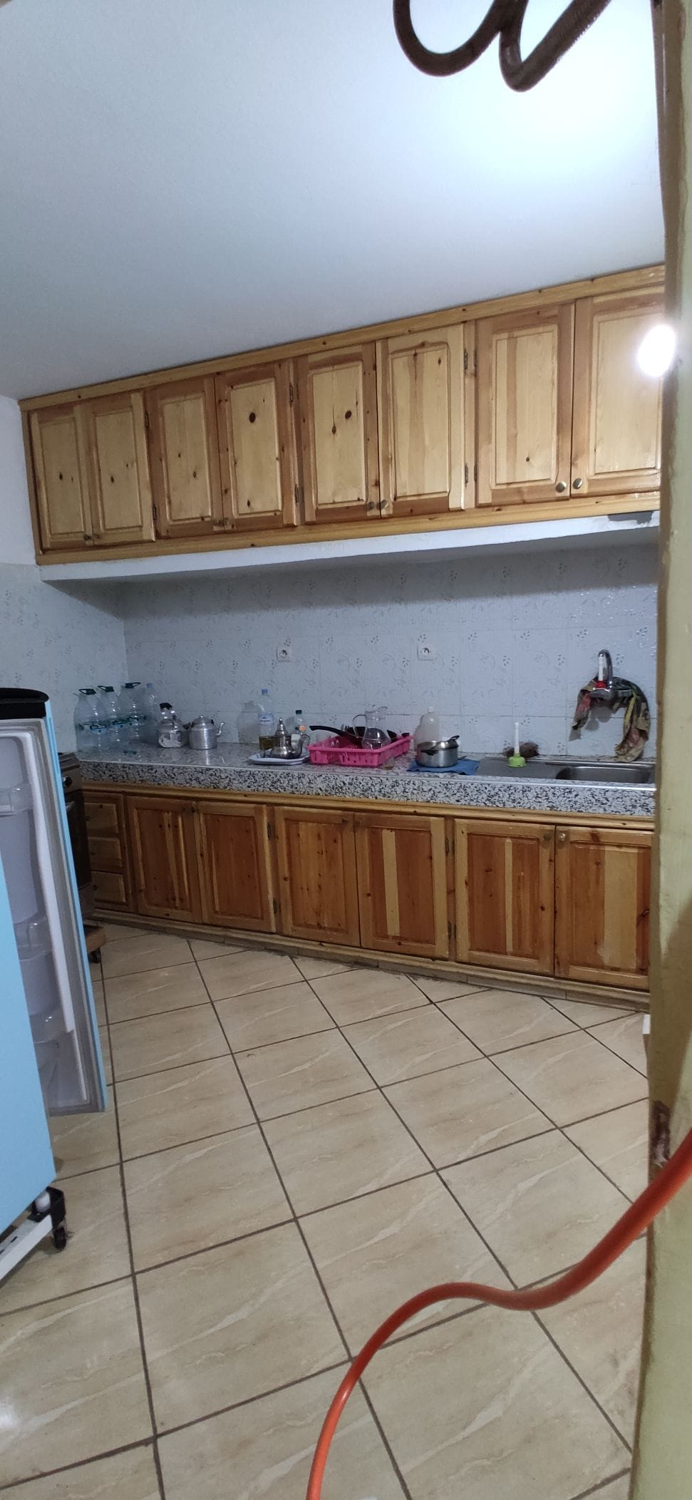 A functional kitchen space features wooden cabinetry above a long countertop with a mix of decorative items and kitchenware. The tiled floor adds a practical touch, while the walls maintain a subtle texture. Appliances, including a refrigerator and a sink, are also visible.