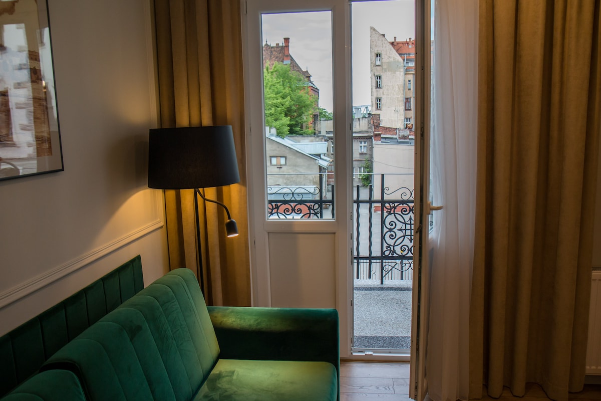 A green velvet sofa is positioned next to a large window, which offers a view of the cityscape. Sheer curtains frame the window, allowing natural light to enter. A modern floor lamp stands nearby, providing additional illumination.