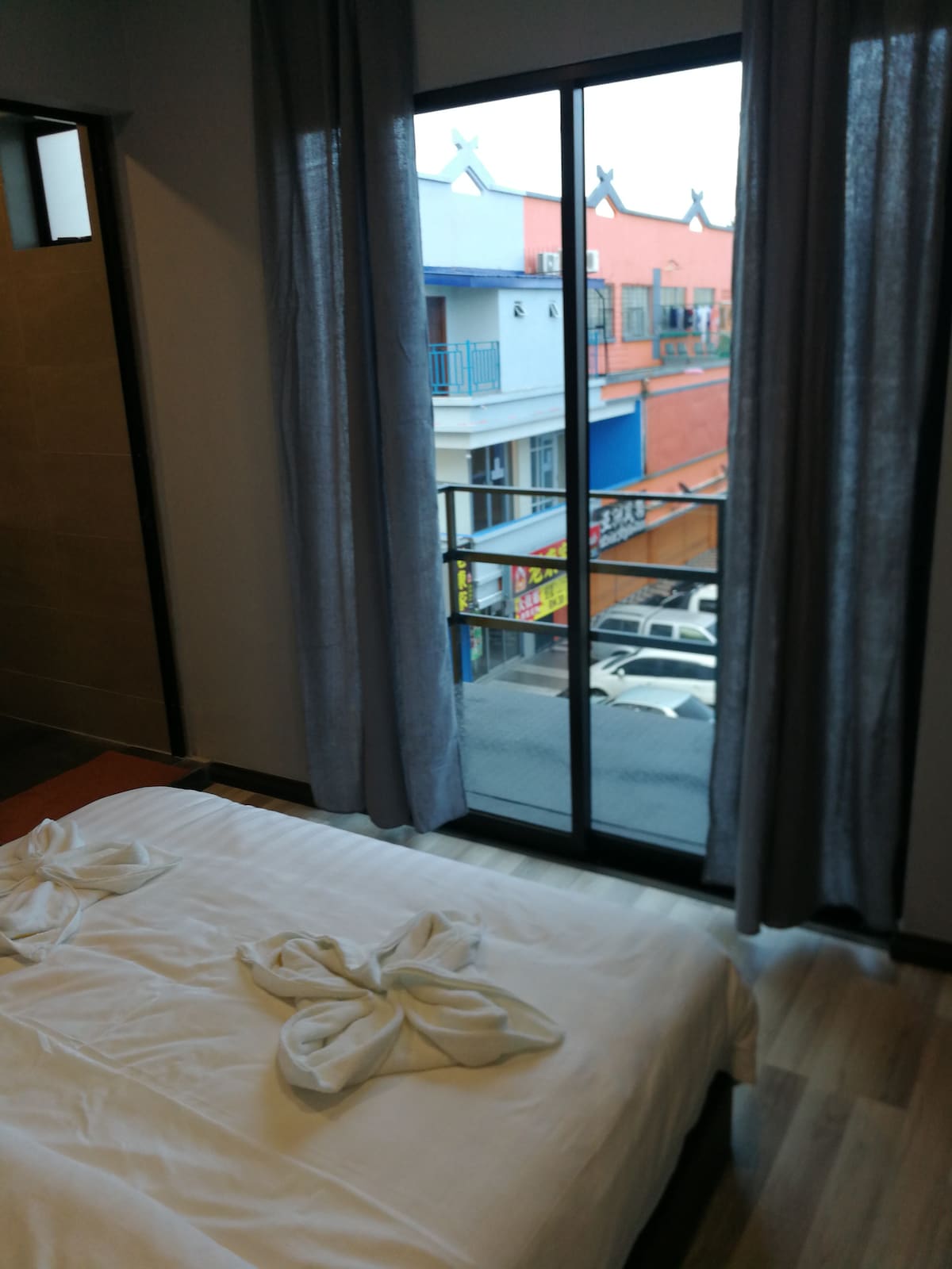 A spacious bed is positioned near a large window, which is framed by airy, light-colored curtains. The view reveals a colorful street scene with buildings and parked vehicles, bringing a sense of activity and local charm to the room.