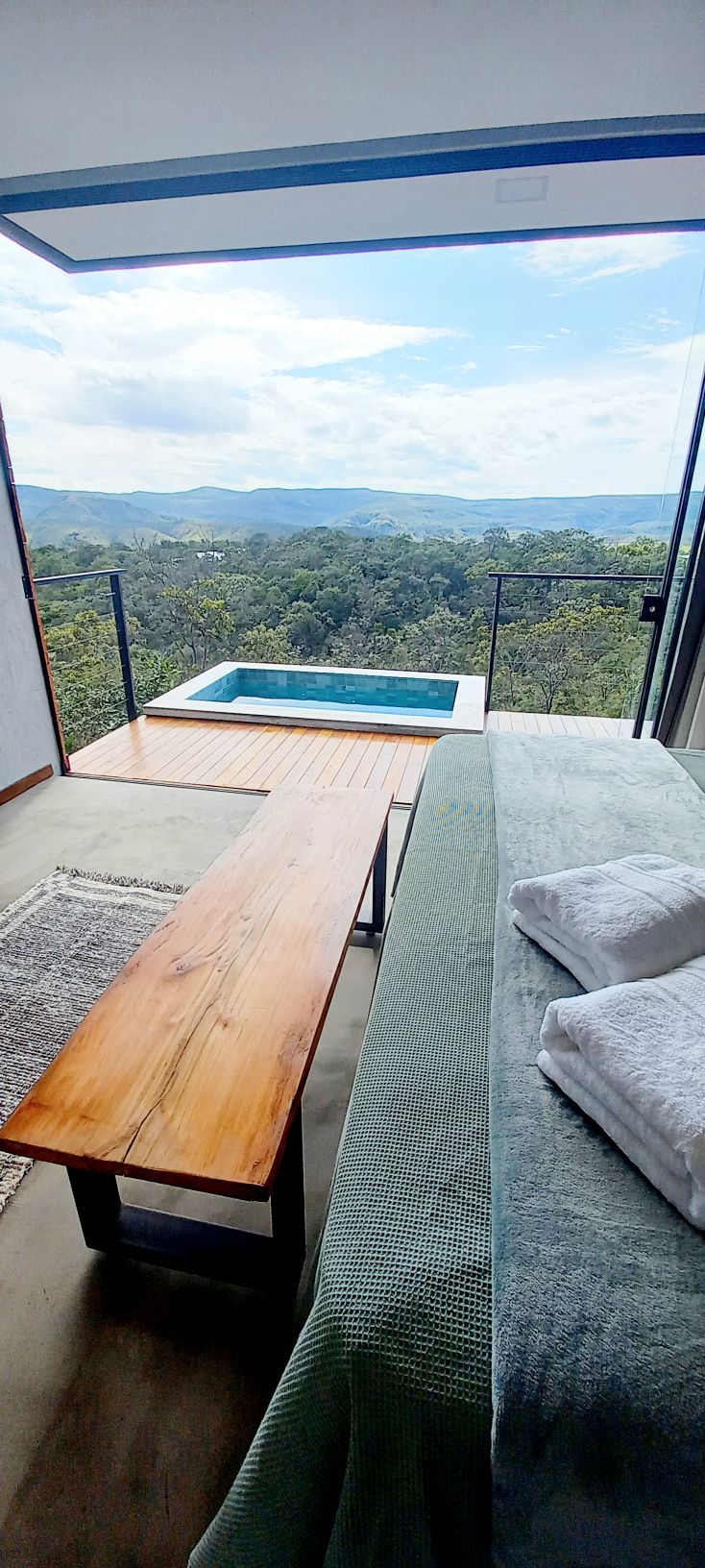 A cozy interior space is presented, featuring a wooden table alongside a spacious bed adorned with plush towels. A small heated pool is visible on the terrace, offering sweeping views of lush green mountains and clear skies beyond.