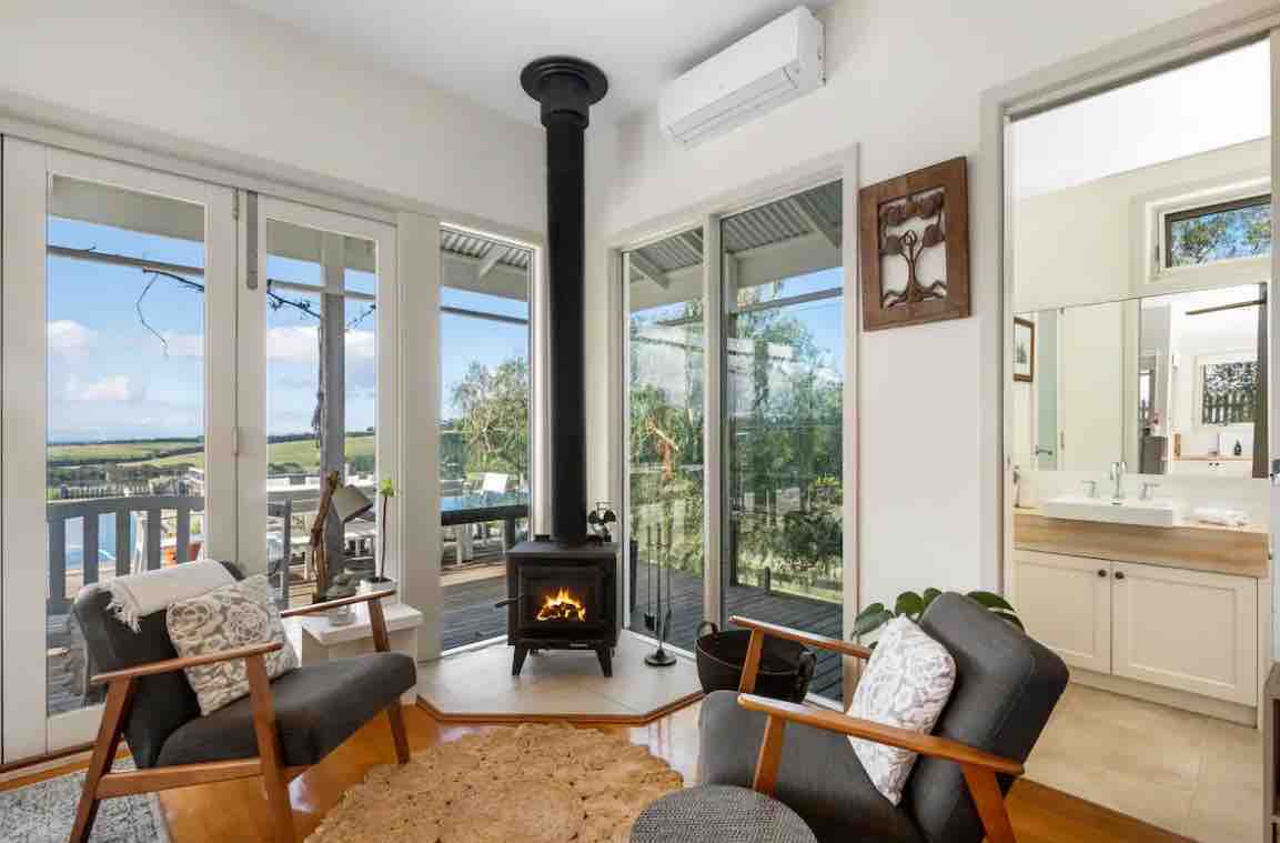 A cozy seating area is presented with two armchairs beside a circular rug. A wood heater stands in front, enhancing warmth. Expansive glass doors provide views of the outdoor deck and surrounding landscape, allowing natural light to fill the space.