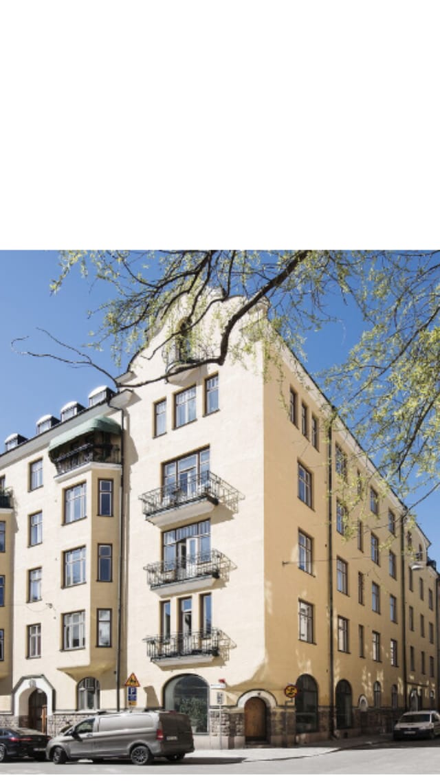 A historic building from 1905 is prominently displayed in this image. The ochre-colored facade features intricate architectural details, including balconies with wrought iron railings and large windows. A tree with fresh foliage provides a natural element against the clear blue sky.