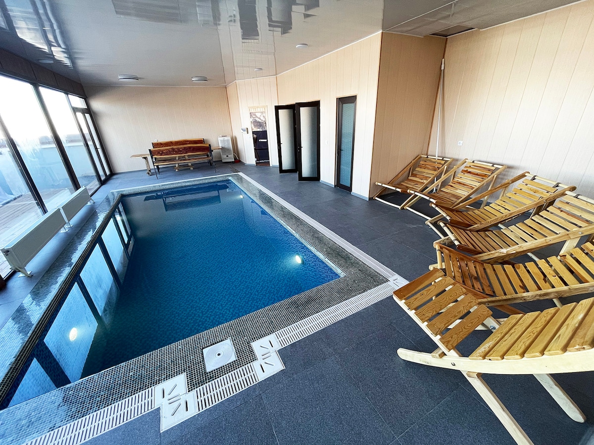 An indoor pool area features a sleek, tiled pool bordered by wooden lounging chairs. Large windows provide ample natural light and panoramic views of the surrounding landscape. Minimalist decor complements the serene atmosphere, ensuring a relaxing environment.