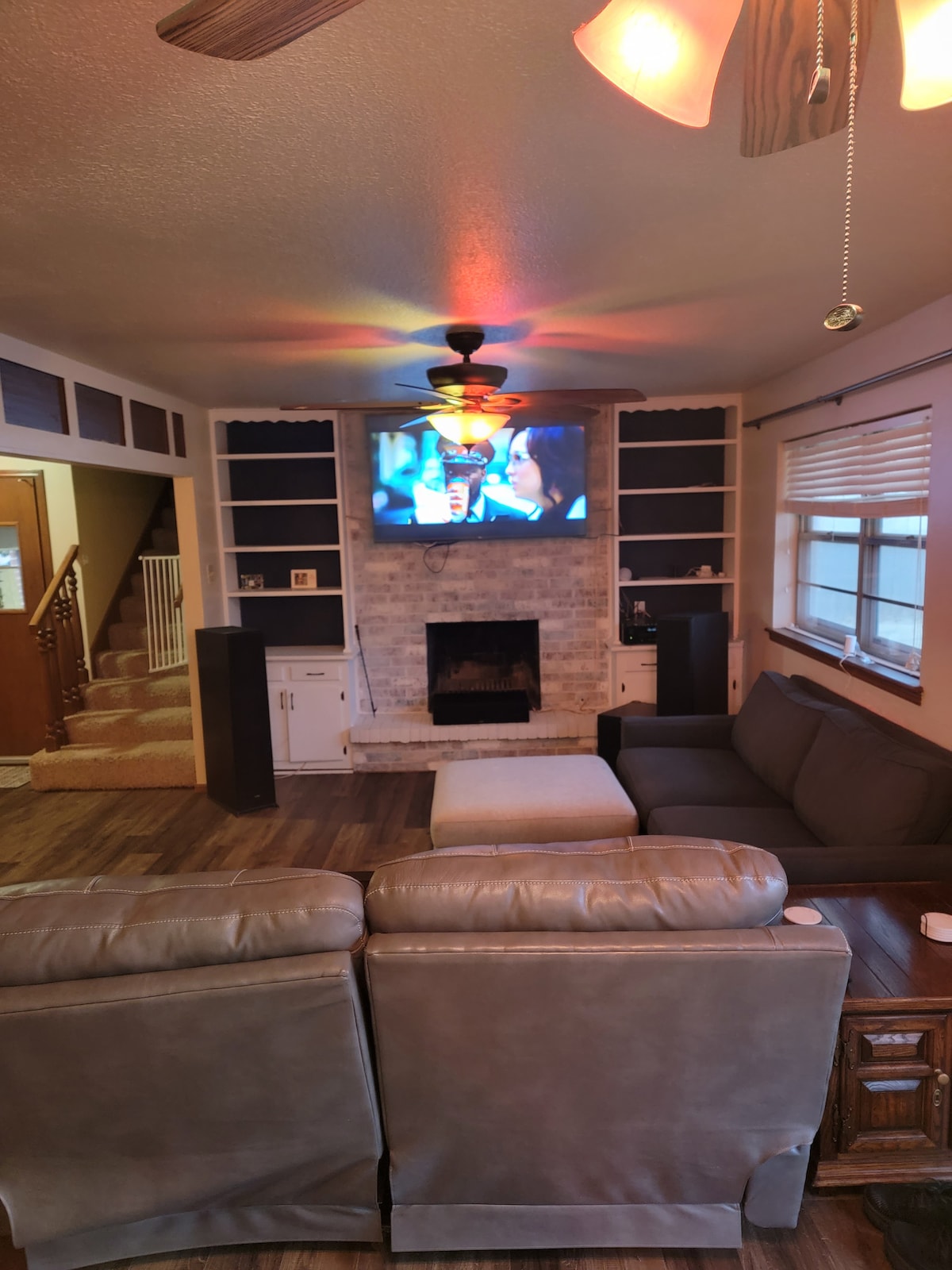 A living room features a comfortable seating arrangement with a large gray sectional couch and an armchair. A fireplace is centered beneath a wall-mounted television. Warm lighting from a ceiling fan casts a cozy ambiance, while built-in shelves flank the entertainment area.
