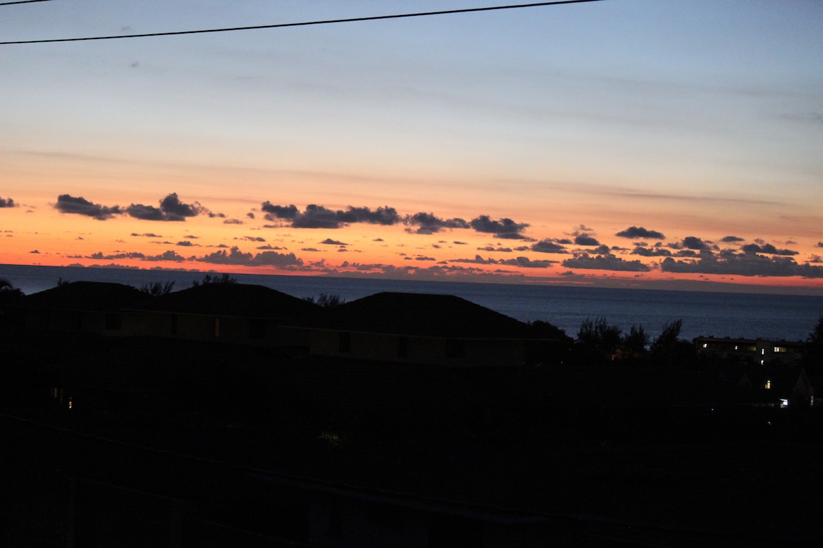 A serene sunset view is captured, showcasing a gradient of colors transitioning from deep orange to soft pink and purple, over the ocean horizon. Silhouettes of distant hills are visible in the foreground, enhancing the tranquil atmosphere of the scene.