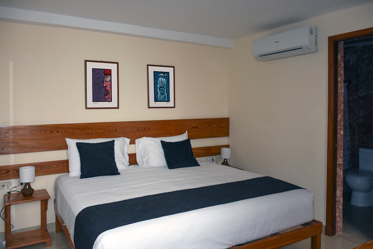 A spacious bedroom features a large bed with dark accent pillows. Two framed artworks adorn the walls above the bed. A bedside table with a lamp is visible, and an air conditioning unit is mounted on the wall, providing comfort during warmer days.