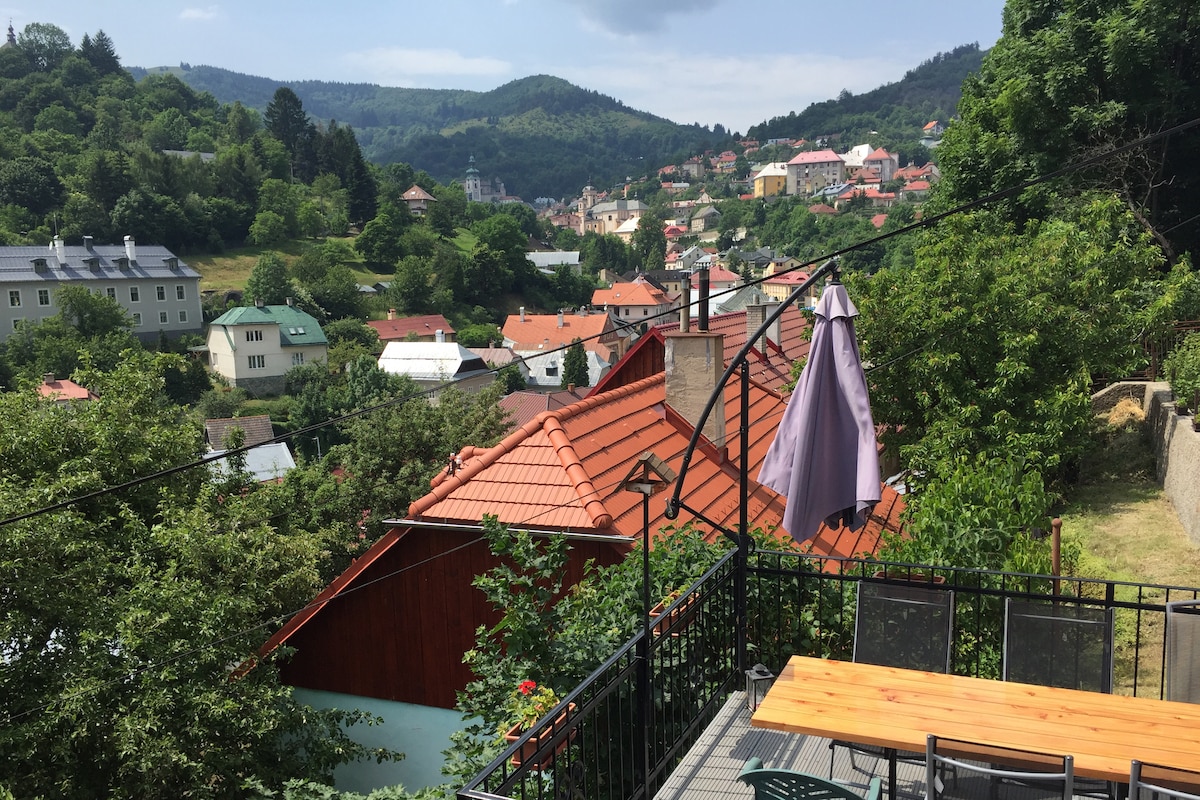 A terrace is seen with a wooden dining table and chairs, providing an expansive view of the surrounding hills and greenery. Various rooftops and homes dot the hillside, creating a picturesque landscape. A large umbrella offers shade for outdoor dining.