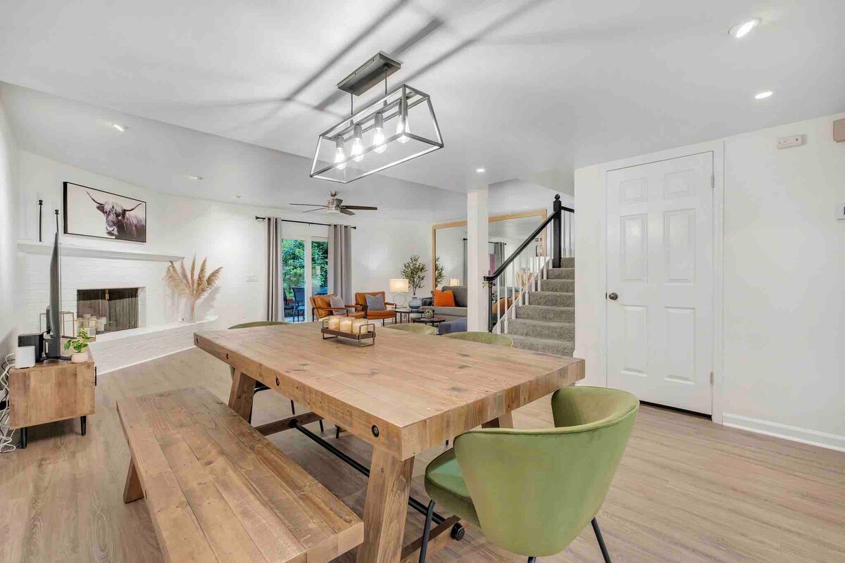 A spacious dining area is highlighted by a large wooden table surrounded by green chairs. A comfortable seating arrangement is visible in the background, with various decorative elements, including wall art and plants, enhancing the inviting atmosphere.