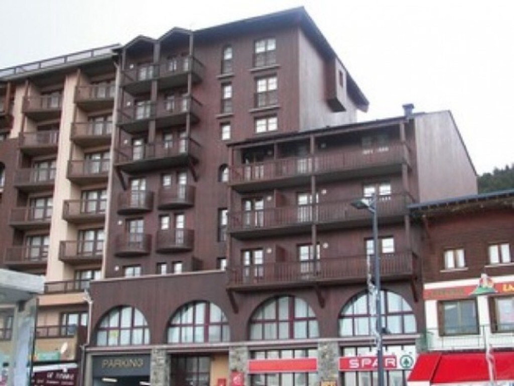 A modern five-story building with a wooden facade is presented. Balconies are visible on the upper floors, and large arched windows are featured on the ground level. Nearby shops are partially seen, contributing to the accessible urban environment.