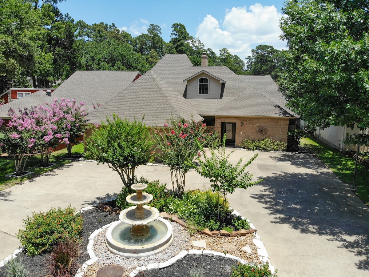 A well-kept exterior of the home is featured, with a landscaped front yard showcasing flowering shrubs and a stone fountain. A paved driveway leads to the front entrance, with the house surrounded by greenery and trees in the background.