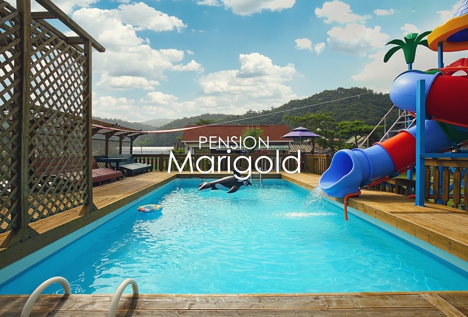 A swimming pool is visible with a bright blue surface reflecting the sky. A water slide is positioned adjacent to the pool, providing added enjoyment for guests. Lounge areas with umbrellas are situated around the pool, surrounded by green hills in the background.