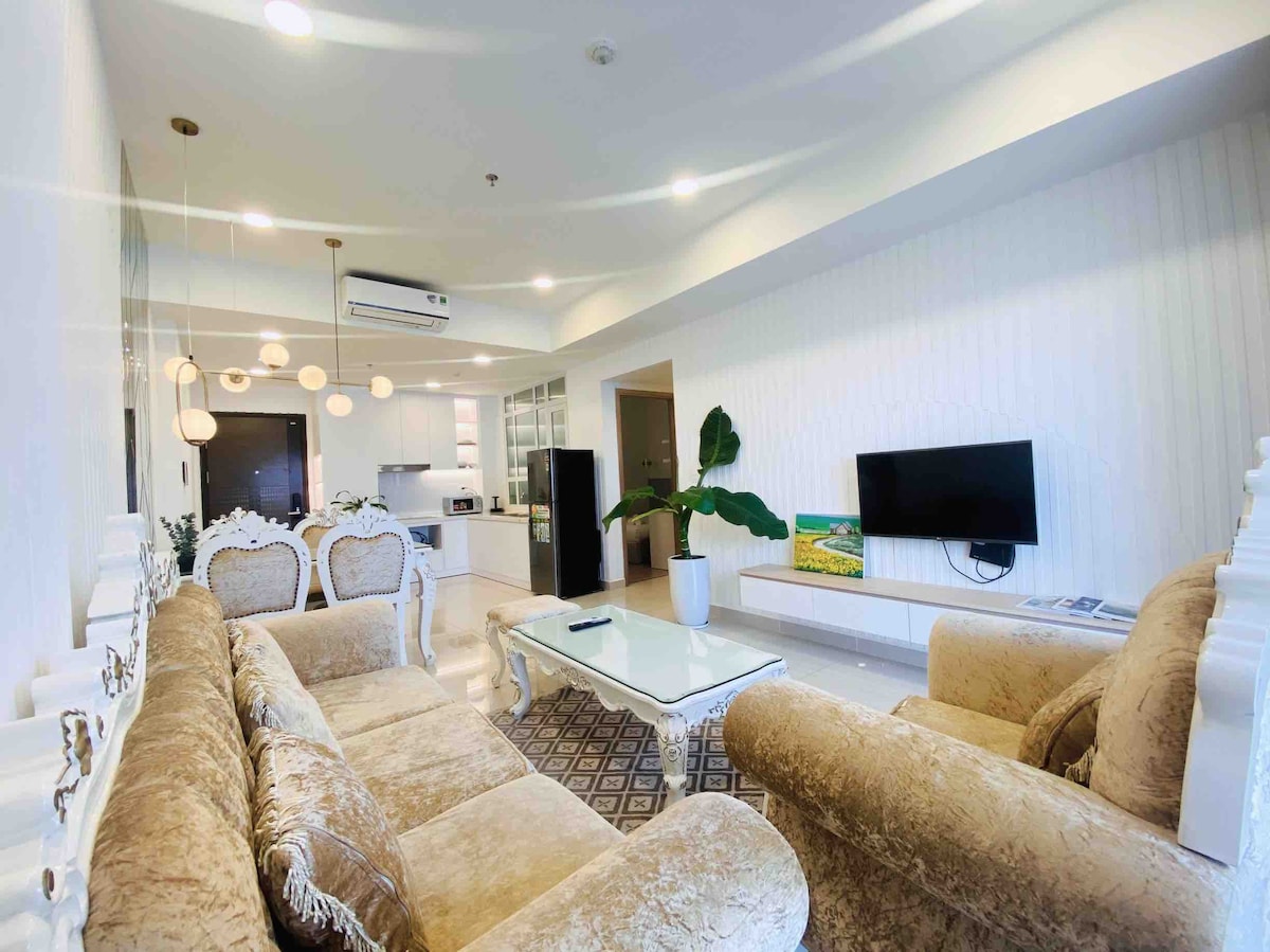 A spacious living area features two decorative sofas with intricate detailing, arranged around a glass coffee table. A television is mounted on the wall, and light fixtures provide illumination. A tall indoor plant adds a touch of greenery, enhancing the overall ambiance.