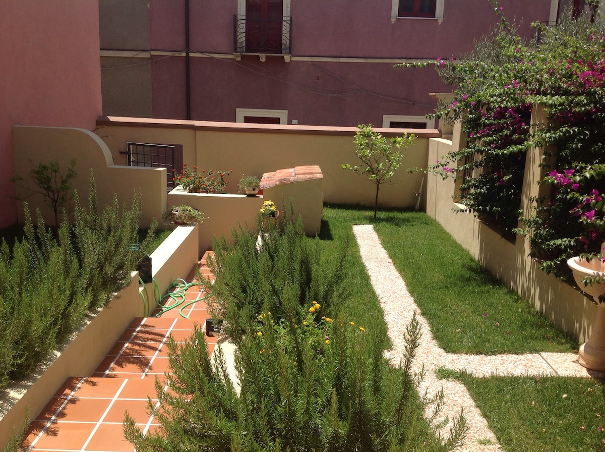 A private garden features abundant greenery and neatly arranged flower beds. A path made of tiles leads through the space, bordered by flourishing plants. A small tree is visible in the background, alongside climbing vines that add texture to the garden walls.