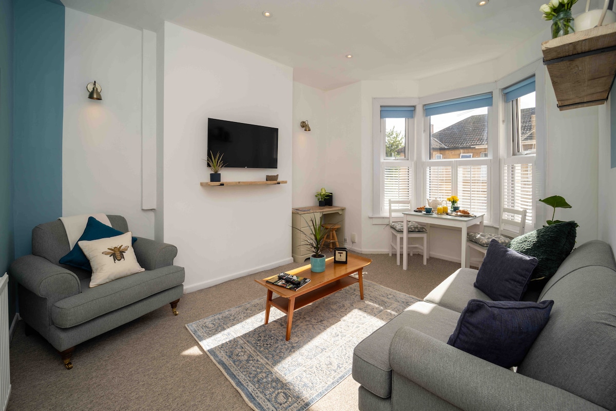 A bright living room features two comfortable sofas facing a mounted television. A wooden coffee table is centered on a decorative area rug. Natural light floods in through large bay windows, illuminating a small dining table set against the wall, enhanced by plants and décor.