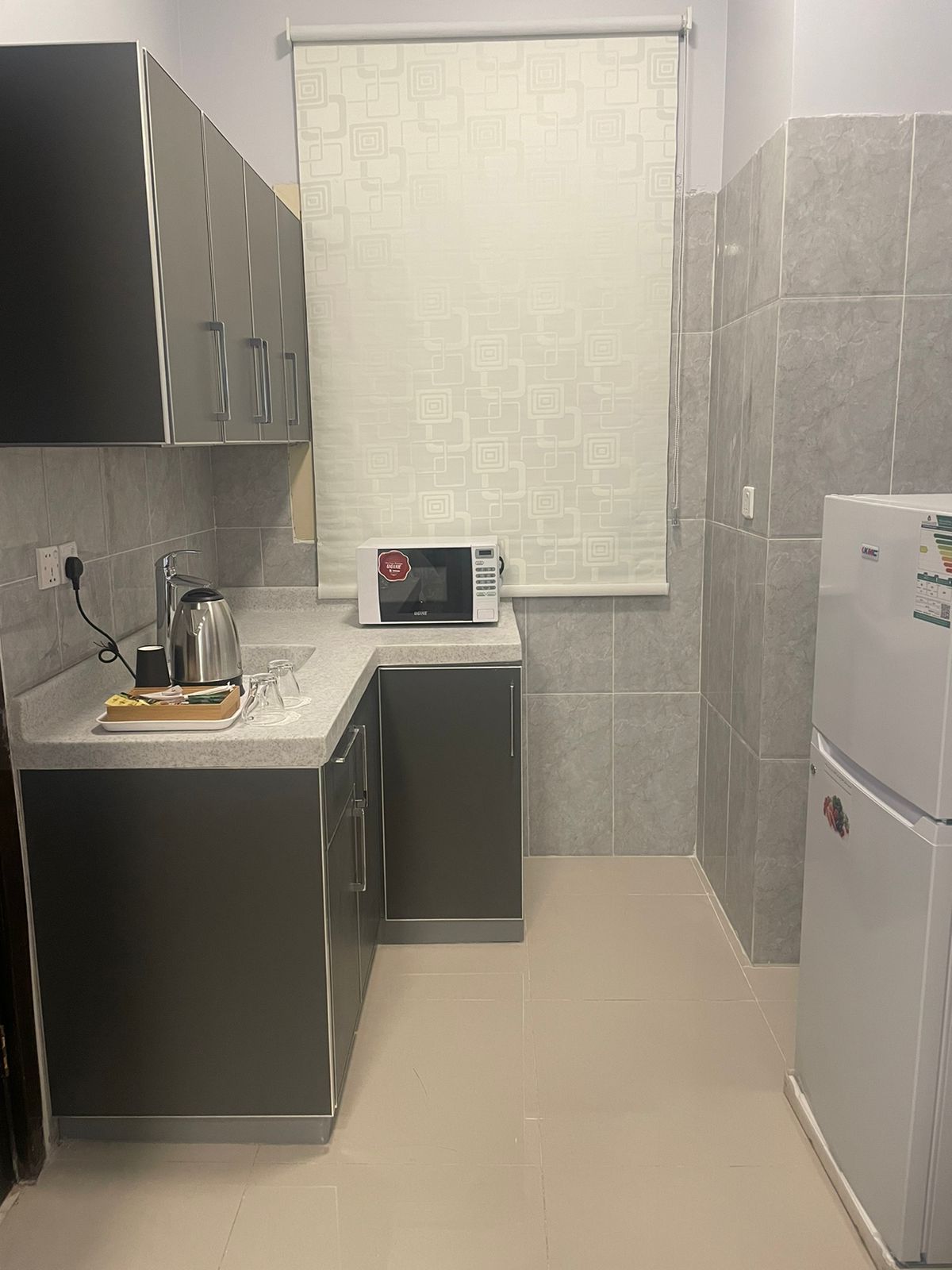 The kitchen features grey cabinetry with a countertop, a kettle, and a microwave on display. A refrigerator stands against the wall, while a window is dressed with a light-filtering shade, allowing natural light to enhance the space.