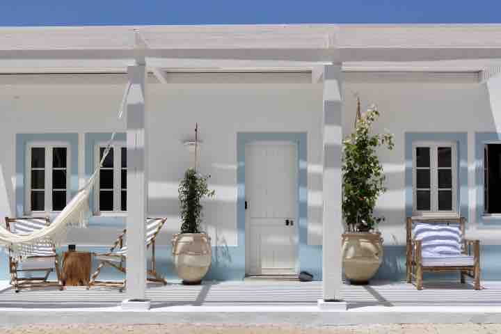 A covered porch features a hammock and two wooden chairs, inviting relaxation. Potted plants add greenery, while white columns and blue accents create a serene atmosphere. Natural light brightens the space, with shadows creating a rhythmic pattern on the flooring.