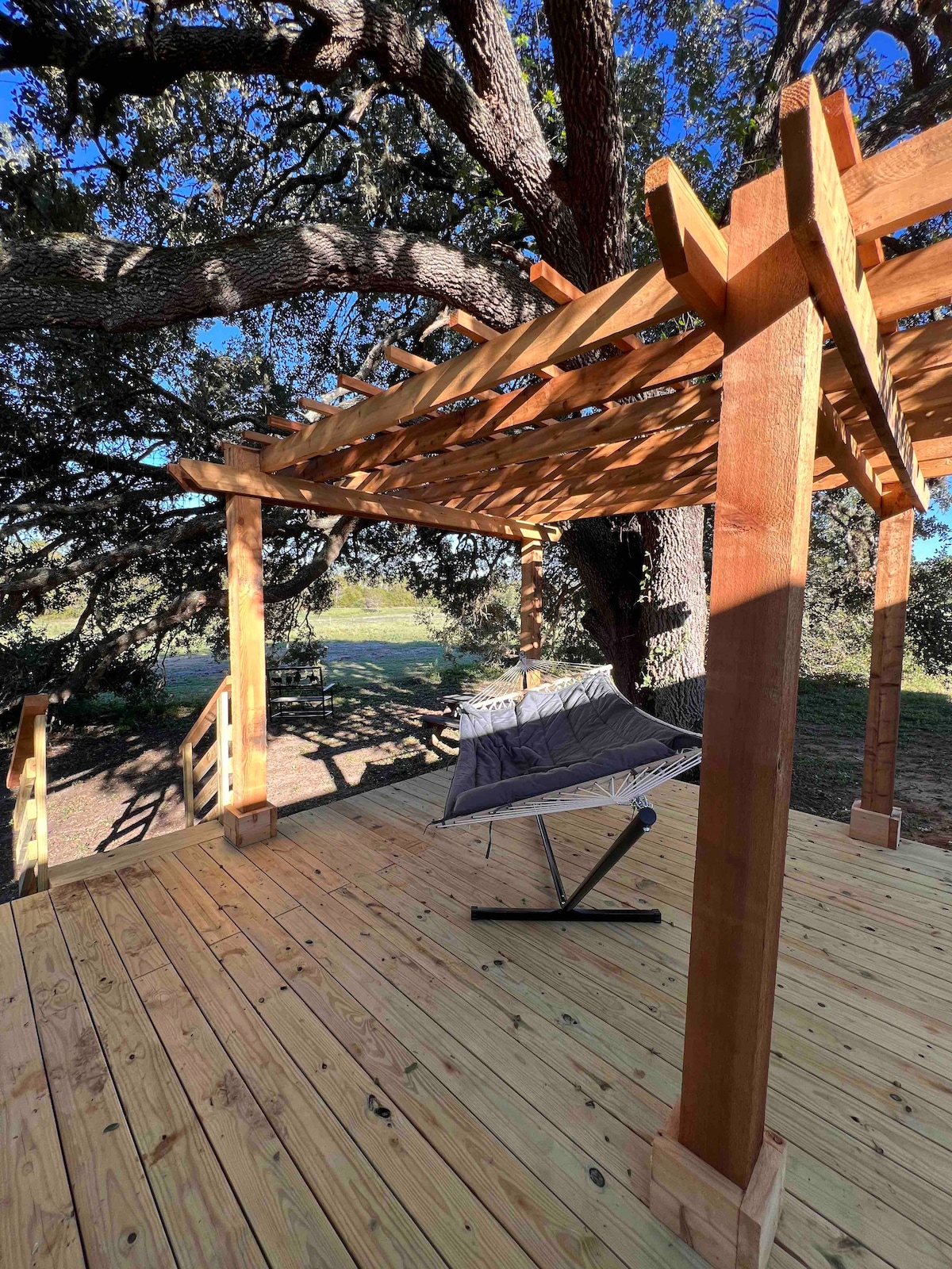 A spacious wooden deck is highlighted under a large oak tree, featuring a sturdy hammock suspended in the center. The deck is surrounded by natural light, with wooden beams and slatted overhang providing partial shade.