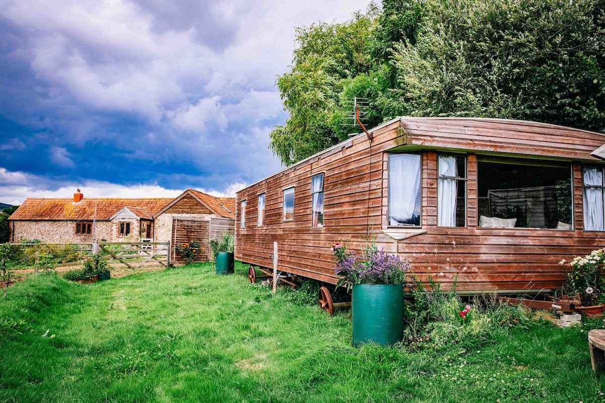 A rustic mobile home made from up-cycled materials is surrounded by lush green grass and flowering plants. The cabin features large windows that allow natural light to enter. In the background, a traditional farm building is visible under a cloudy sky.