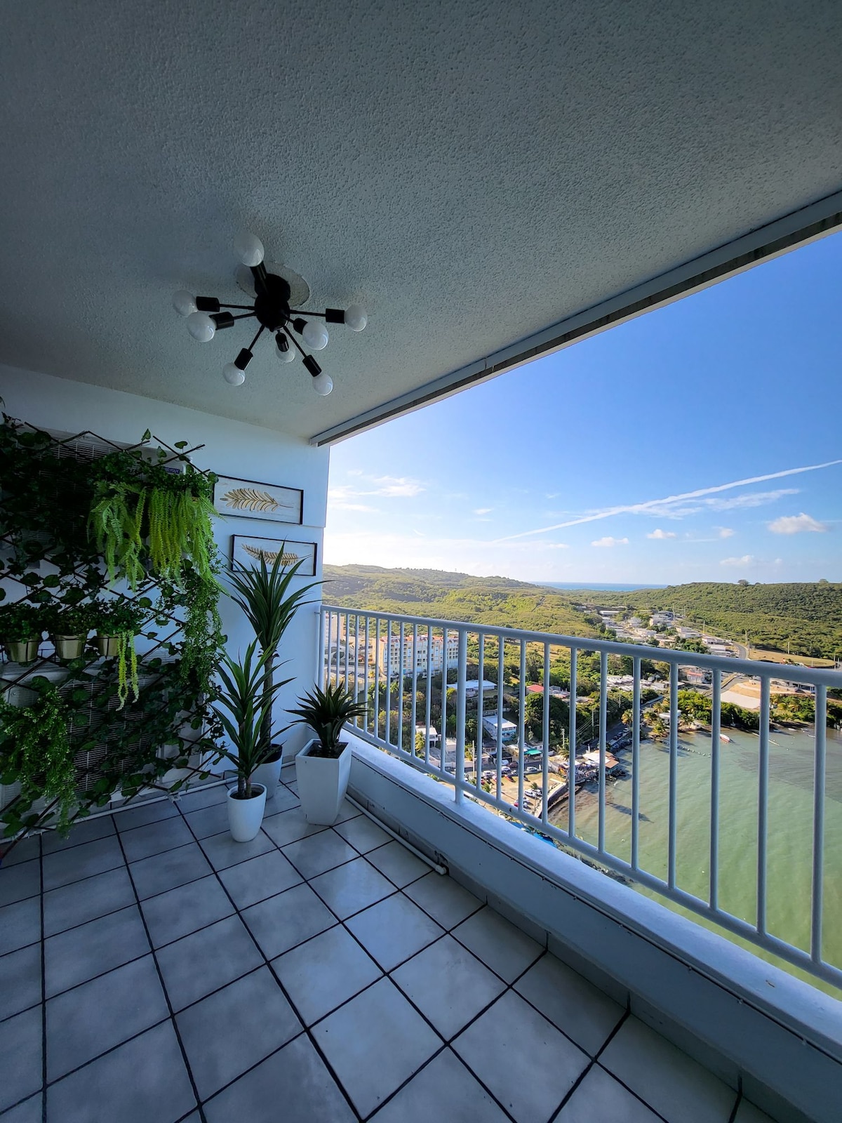 A balcony space is revealed, featuring a striking view of the surrounding hills and water. Potted plants add greenery to the scene, while a stylish light fixture hangs from the ceiling. Spaces of tiled flooring enhance the open feel of the area.