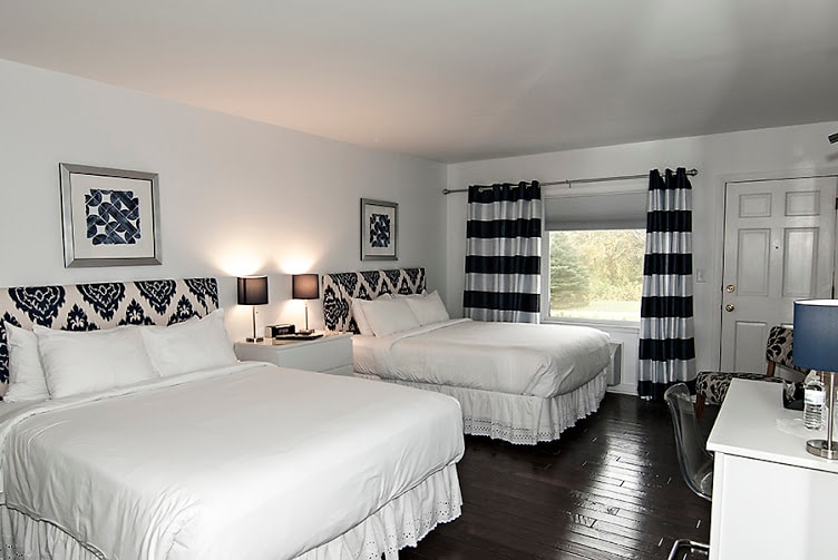 A well-appointed guest room features two spacious beds with patterned headboards. Light-colored bedding contrasts with dark hardwood floors. A window with striped curtains allows natural light to enter, and a desk with a lamp is positioned nearby, enhancing the room's functionality.