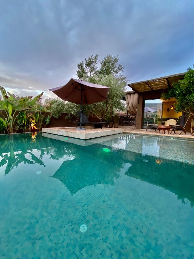 A tranquil pool area is surrounded by lush greenery, featuring a large umbrella over a seating space. The water reflects the surrounding landscape, while a covered area with chairs and a table is visible in the background.