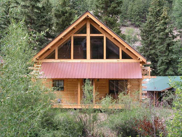 A log cabin exterior is displayed, showcasing a steeply pitched roof with a metal awning. Large windows allow natural light to fill the interior, framed by dense greenery. The surrounding forest adds a natural touch to the cabin's rustic charm.