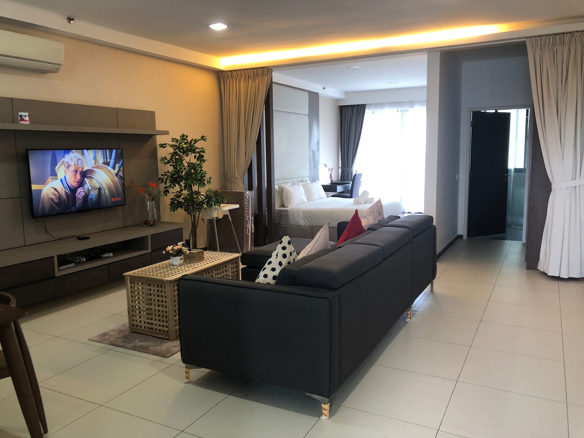 A spacious studio unit is displayed, featuring a modern seating area with a dark sofa and decorative pillows. A sleek entertainment unit holds a television, with a dining table nearby. Natural light fills the room from large windows, leading to a well-appointed sleeping area.