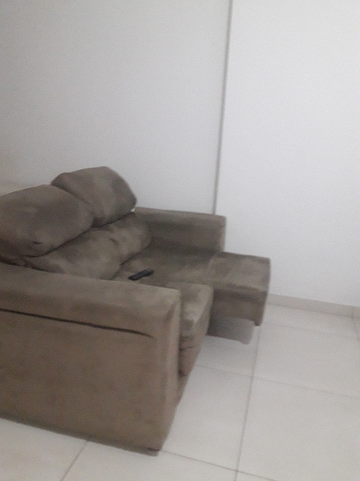 A comfortable armchair is positioned against a plain white wall. The chair features a soft fabric in a neutral tone, with a remote control resting on the seat. The flooring is tiled, contributing to the room's clean and simple aesthetic.