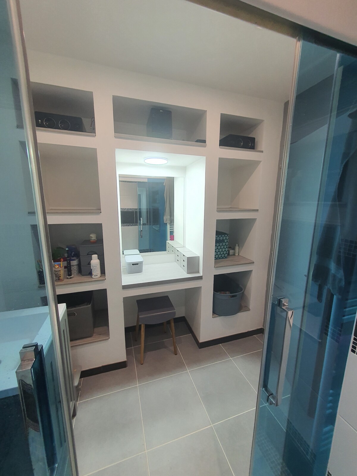 A modern bathroom features a built-in vanity area surrounded by shelves. A stool is positioned in front of the well-lit mirror, with various toiletries neatly arranged on the shelves. The space showcases a glass shower enclosure and is designed with light-colored tiles.