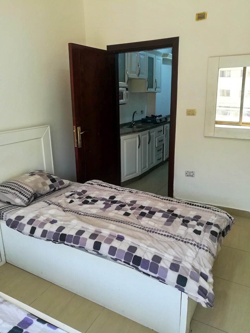A well-lit sleeping area features a single bed with a patterned comforter in shades of white and purple. A wooden door opens to a view of a kitchenette, while a window allows natural light to enter the space.