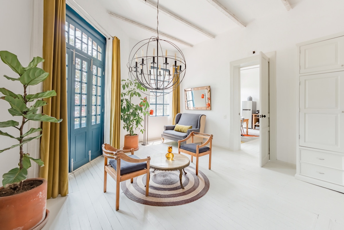 A bright living area is adorned with large windows framed by mustard-yellow curtains. A circular coffee table sits on a patterned rug, surrounded by two blue accent chairs and two wooden armchairs. A potted plant adds a touch of greenery to the space.