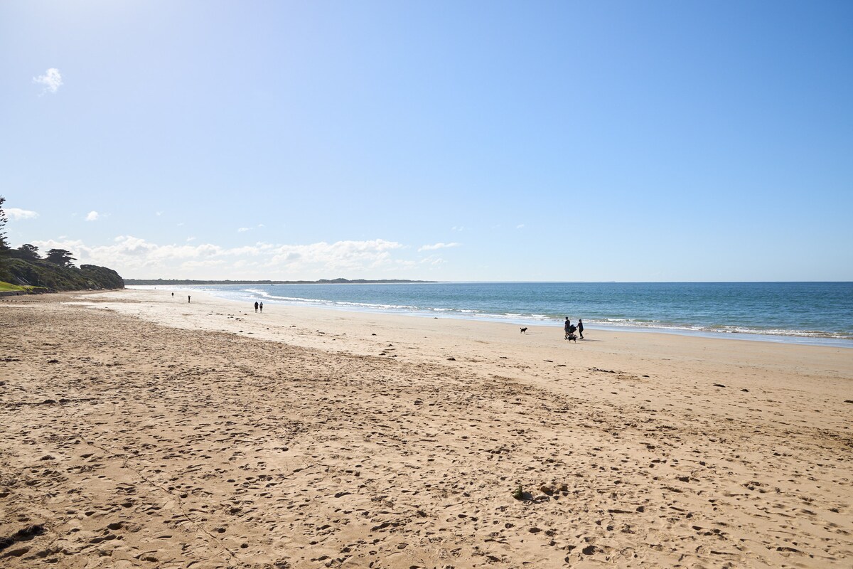 A sandy beach stretches along the shoreline, meeting the gentle waves of the sea. People can be seen walking along the beach, enjoying the open space and clear blue sky. The scene is tranquil, with distant greenery visible along the beach's edge.