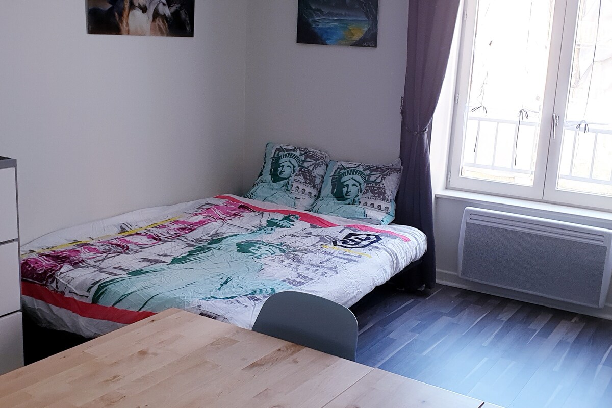 A cozy sleeping area is presented with a spacious bed featuring a colorful blanket. Natural light enters through a large window dressed with sheer curtains. A simple wooden table stands nearby, contributing to the functional layout of the studio.
