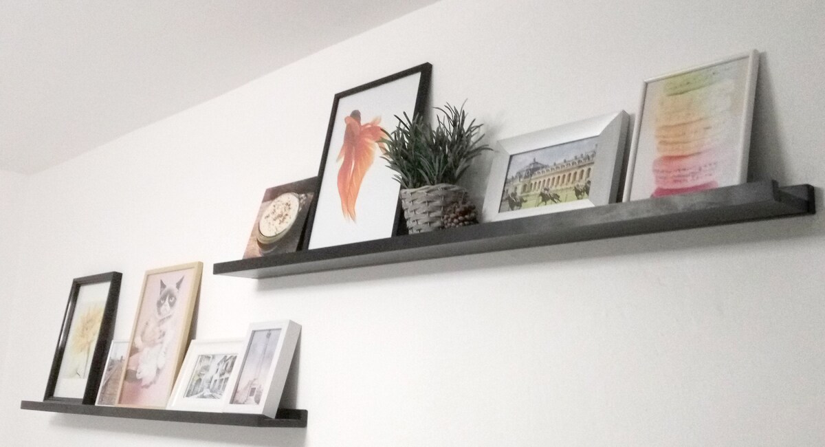 Two dark wooden shelves adorn a white wall, displaying various framed artworks and photographs along with a small decorative plant in a woven pot. The collection features a mix of colors and styles, adding a touch of personality to the space.